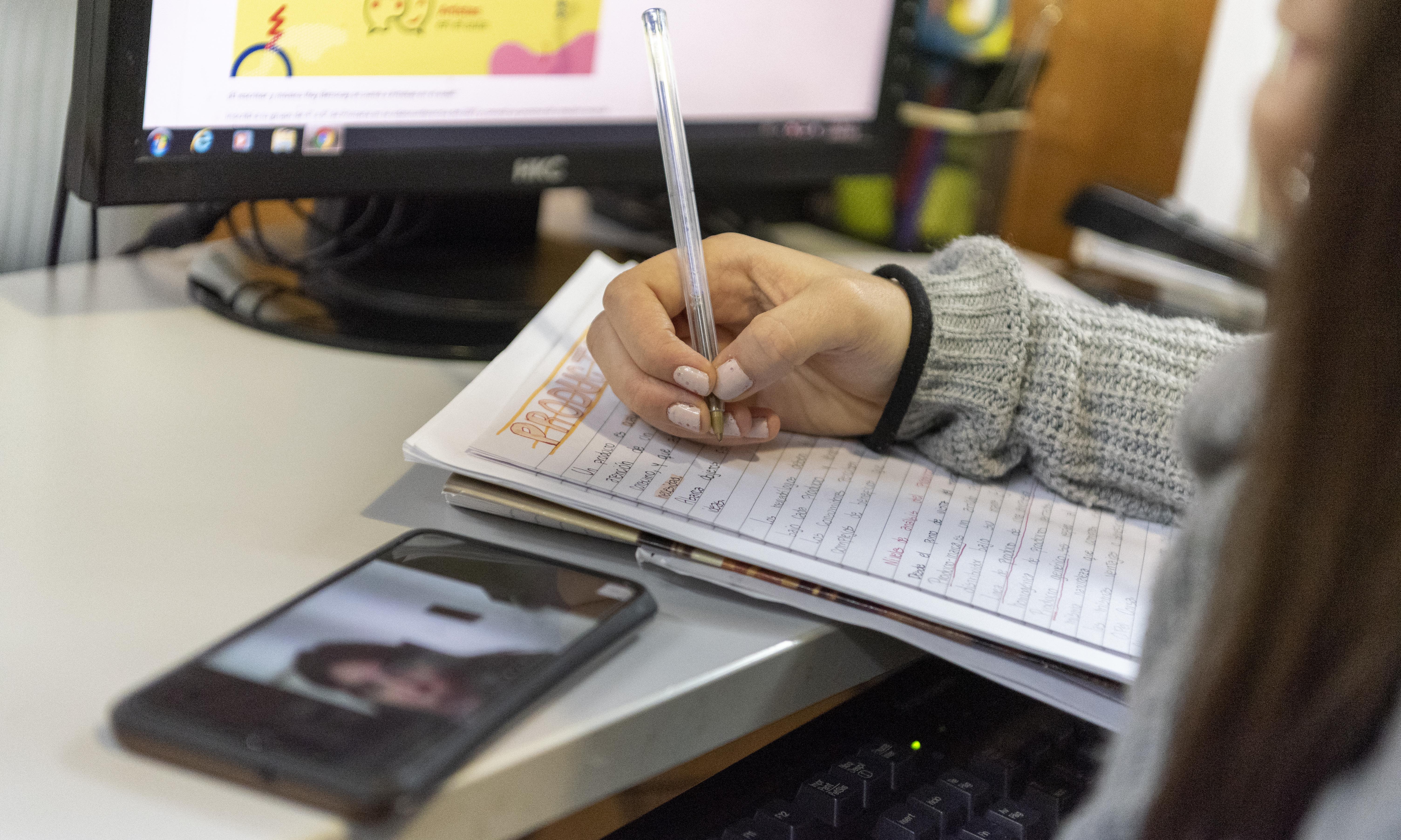 Persona estudiando a distancia en un escritorio con computadora, teléfono celular y cuadernola.